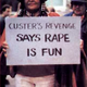 A woman wearing Native American signifiers holds a protest sign that says "Custer's Revenge says rape is fun".